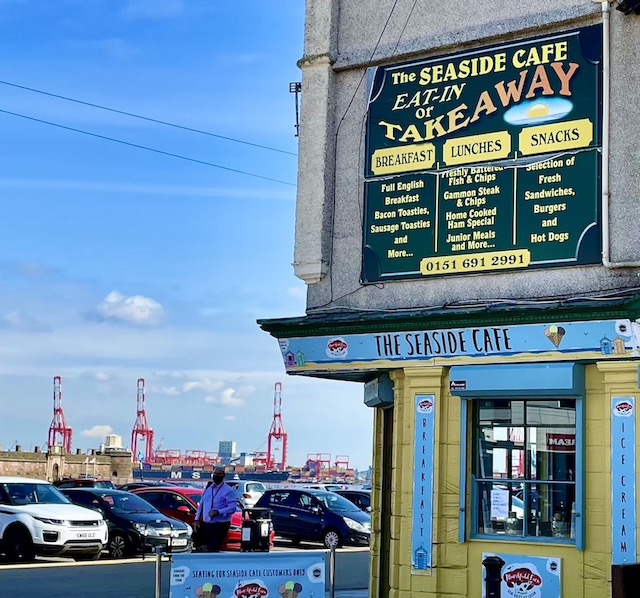 The seaside cafe, with cars parked in the near side, and dock cranes in the distance