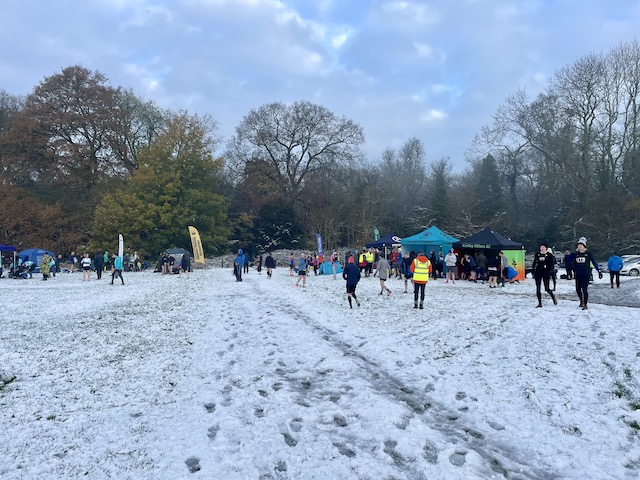 Snowy field with lots of footprints. In distance, tents and banners with people milling around. And trees behind that.