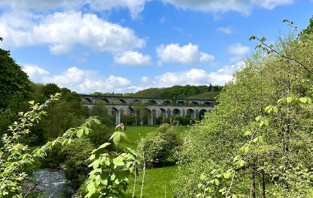 Aqueduct in background with trees in foreground and river in left foreground. Blue skies with light white clouds 