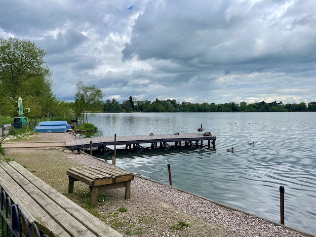 A mere with a small pier with a couple of overturned boats and ducks in the water surrounded by trees and a couple of benches