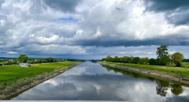 River stretching into the distance with  grassy edges and trees edging them. Dramatic cumulonimbus clouds above being reflected in the water.