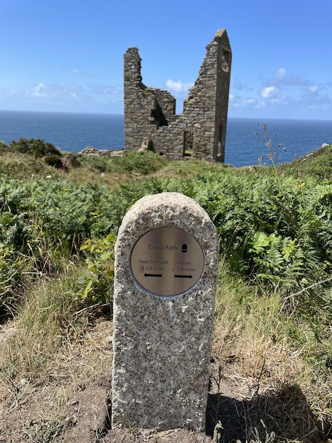 A sign for the coast path in the foreground with another engine room ruin in the background with the sea and sky behind that.