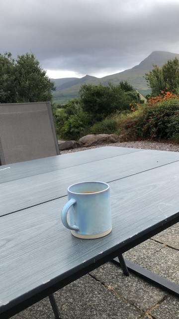 A mug of tea on an outdoor table with the mountain views and green trees in the background.