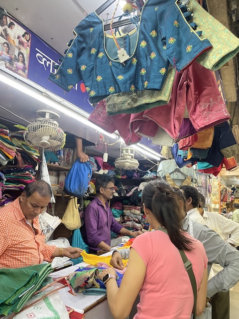 A bustling kiosk with blouses hanging up, a woman talking to a man and several other men organising the sewing. A hand is passing a bag down from a trap door above them.