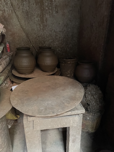 A stool with a flat disc of a potter's wheel on it, with some ready thrown pots behind it in a room.