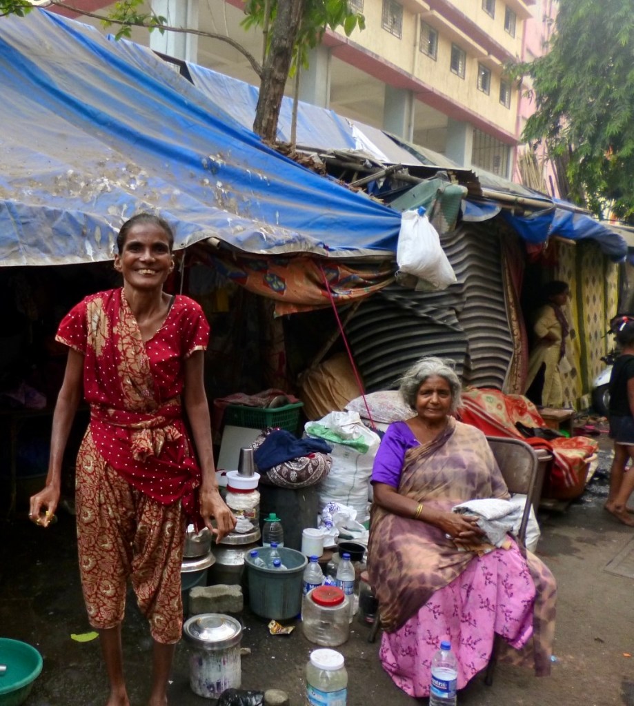 Two women by some tarpaulin tents standing and sitting with their pots and possessions.