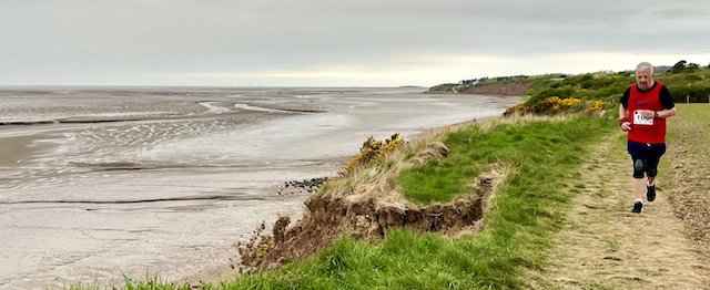 A man in a red vest running along a trail path parallel to the beach on his right.
