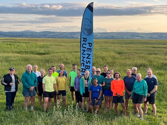 A lot of runners on a long grassy ground with the Pensby Runners flag in the middle, with clouds and hills in the background. 