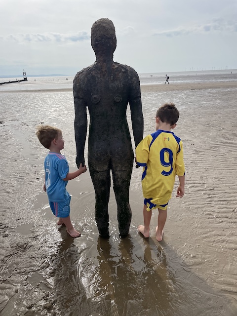 Two boys holding hands with one of the iron men on Crosby beach