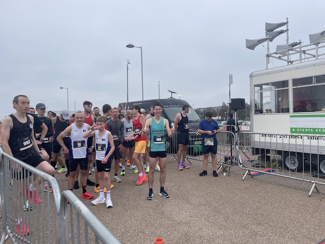 Several runners standing by metal barriers waiting for the race to start, and our Ben Taylor with his thumbs up to me.