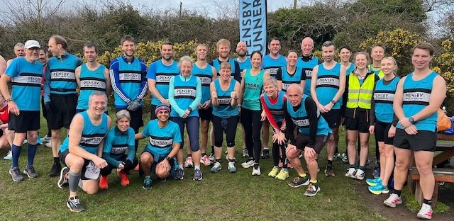 A big crowd of runners for Pensby with the club flag behind, posing outside on the grass