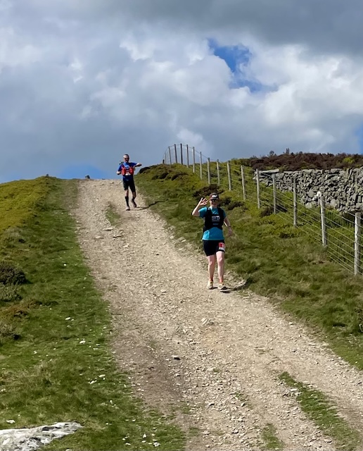 Two runners running down a gravel path with grass verges, the front one, from Pensby Runners, waving.