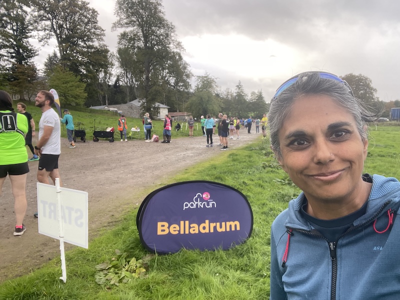 Runners in the background on grass and a muddy path. The Parkrun 'Belladrum' sign in the foreground with me doing a selfie.